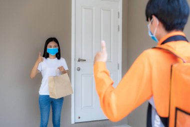 Asyalı bir kadın yemek paketini kapı tokmağından alır ve başparmağıyla temas kurar. Ya da bisikletli bir teslimatçıyla temas kurmaz. Enfeksiyon riskine karşı sosyal mesafe için eve bisikletle gelir..