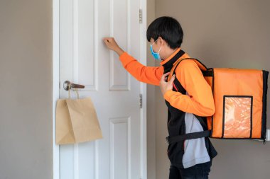 Asyalı teslimatçı, enfeksiyon riskine karşı sosyal mesafe koymak için evdeki teslimatçıdan ya da teslimatçıdan uzak kalmak için kapı koluna yiyecek paketi gönderir..