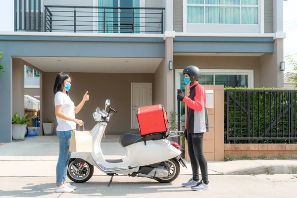Asian woman pick up delivery food bag from box and thumb up form contactless or contact free from delivery rider with bicycle in front house for social distancing for infection risk.