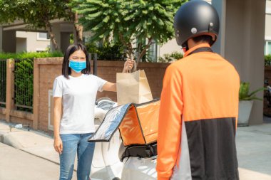 Asyalı bir kadın kontak kurmak için kutudan yiyecek torbası alıyor ya da enfeksiyon riskine karşı sosyal mesafe için evin önünde bisikletli bir teslimatçıyla temas kurmuyor..