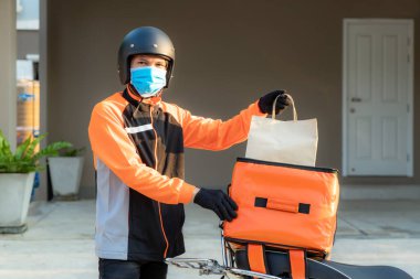 Teslimatçı Asyalı adam turuncu üniformalı koruyucu maske takıyor ve yiyecek paketini scooter 'lı çanta, ekspres yemek teslimatı ve online alışveriş konseptiyle müşteri konutlarının önüne göndermeye hazır.