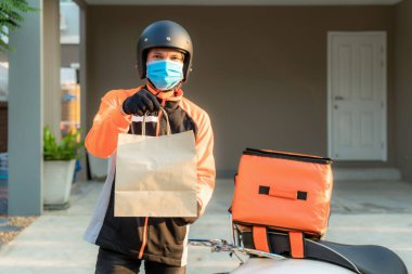 Teslimatçı Asyalı adam turuncu üniformalı koruyucu maske takıyor ve yiyecek paketini scooter 'lı çanta, ekspres yemek teslimatı ve online alışveriş konseptiyle müşteri konutlarının önüne göndermeye hazır.