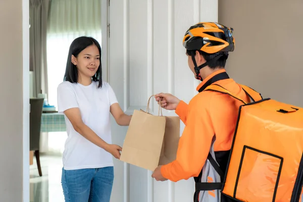 Asian man courier on bicycle delivering food in orange uniform smile and holding food bag in front house and Asian woman accepting a delivery of boxes from deliveryman.