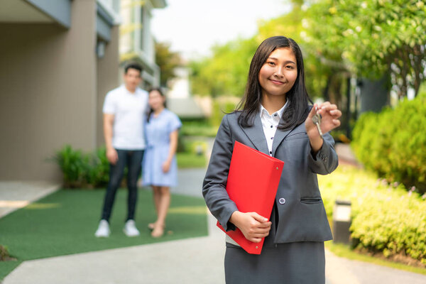Asian real estate agent or realtor woman smiling and holding red file with showing house key with happy Asian couple hugging in front of thier house.