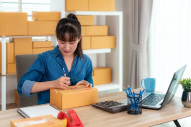 Asyalı genç iş kadını evde online alışveriş için çalışıyor, posta kutusunda ofis ekipmanlarıyla birlikte adres yazıyor, girişimci yaşam tarzı kavramı