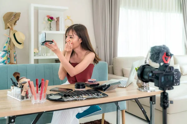 Asian woman vlogger present or teaching make up beauty cosmetic product on her face and broadcast live video to social network by internet at home, beauty blogger concept.