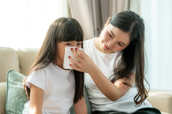Portrait of cut Asian girl blowing snot into the napkin with her mother is keeping it near her nose with care. Visuals of people feeling sick and suffering from common ailments such as the flu, headaches.