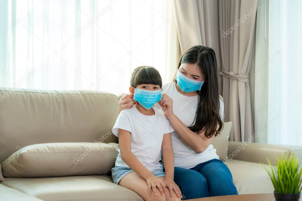 Asian mother wearing wearing to her daughter healthy face mask sitting in living room at home to prevent PM2.5 dust, smog, air pollution and COVID-19. Healthcare concept.