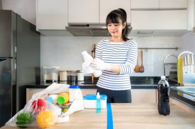 Asian young woman laying out groceries on a divided table and wiping down yogurt or milk containers bottle with disinfectant take to eliminate the chances of contamination COVID-19. clipart
