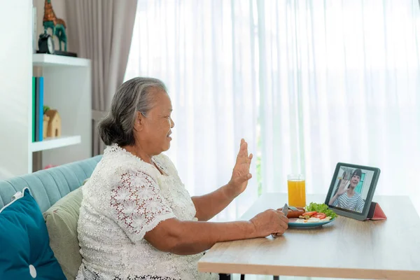 Asian elderly woman virtual happy hour meeting and eating food online together with her daughter in video conference with digital tablet for a online meeting in video call for social distancing