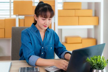 Asyalı iş kadını internet alışverişi için evde çalışıyor, bilgisayarını ofis ekipmanlarıyla ve girişimci yaşam tarzı konseptiyle müşterisiyle sohbet ediyor.