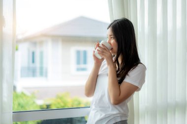 Güzel Asyalı kadın yatağında dinlenmiş olarak uyanıyor ve pencerenin kenarındaki perdeleri açıyor ve sabah yatak odasındaki yatak odasında temiz hava almak için bir fincan kahveyle pencereden dışarı bakıyor.