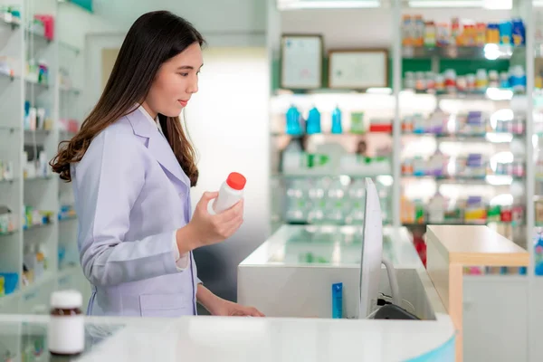 Confident Asian young female pharmacist with smile holding a medicine bottle and searching that product in the computer database in the pharmacy drugstore. Medicine, pharmaceutics, health care