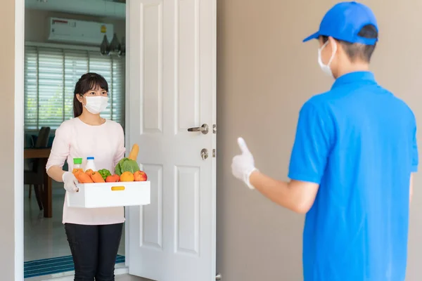 Asian woman pick up groceries box of food, fruit, vegetable and drink and thumb up form contactless or contact free from delivery man in front house for social distancing for infection risk.