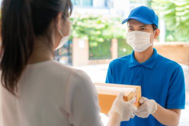 Yüz maskesi ve mavi üniformalı Asyalı kurye evin önünde karton kutular tutuyor ve kadın COVID-19 salgını sırasında teslimatçıdan gelen kutuları kabul ediyor..