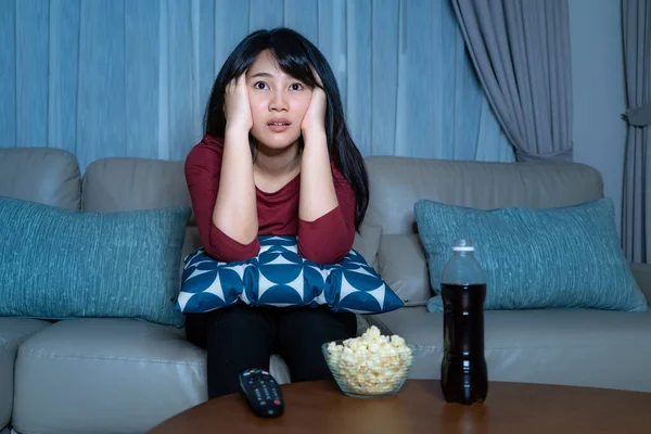 Young asian woman watching television suspense movie or news looking shocked and excited eating popcorn late night at home living room couch during time of home isolation