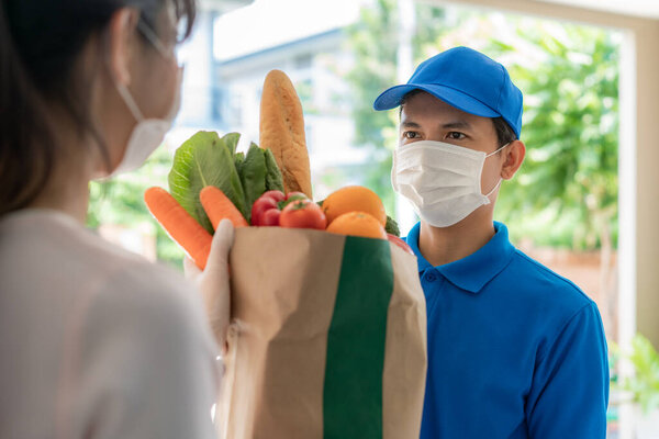 Asian delivery man wearing face mask and glove with groceries bag of food, fruit, vegetable give to woman costumer in front of the house during time of home isolation.