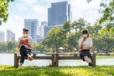 Asyalı genç adam ve kadın kitap okuyor ve akıllı telefondan sohbet ediyor ve maske takıyor. 2 metre uzaklıktalar. Enfeksiyon riskine karşı sosyal mesafe için COVID-19 virüslerinden korunuyorlar.