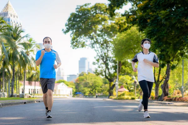 Asian young couple woman and man are jogging and exciseing outdoor in city park and wearing protective mask on face for stay in fit during Covid-19 pandemic in Bangkok, Thailand