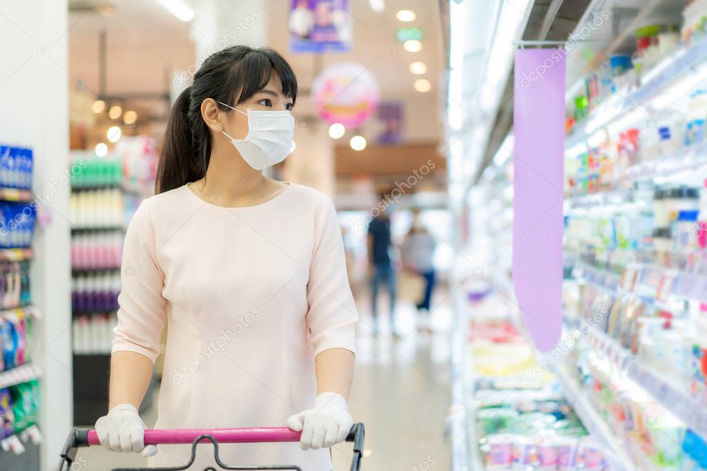 Asian woman with hygienic mask and rubber glove with shopping cart in grocery and looking for yogurt or daily fresh milk to buy during covid-19 outbreak for preparation for a pandemic quarantine