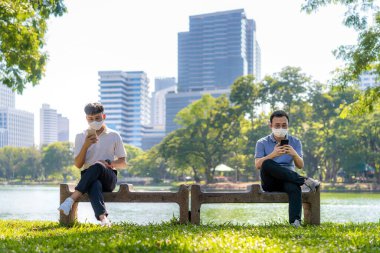 Akıllı telefonda sohbet eden ve maske takan iki Asyalı genç adam 2 metre uzaktan oturarak enfeksiyon riskine karşı sosyal mesafeyi korumak için COVID-19 virüslerinden korunuyor.