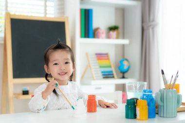 Asyalı anaokulu öğrencisi kız kameraya bakıp gülümserken evdeki oturma odasına akrilik boyayla plastik bebek boyası çiziyor. Evde eğitim ve uzak öğrenim