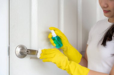 Asyalı bir kadın şişeden mavi bir dezenfektan sıkarak kapı kollarını dezenfekte ediyor. Virüsü ve bakterileri önle, kovid19, koronavirüsü, alkol temizleyiciyi önle. Evde hijyen kavramı.