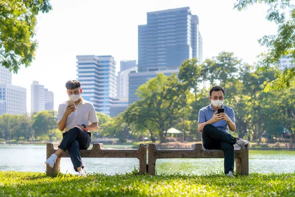 Two Asian young man chatting in smartphone and wearing mask sitting distance of 6 feet distance protect from COVID-19 viruses for social distancing for infection risk at par