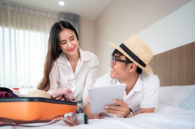 Mutlu çift yatak odasındaki bavullarını topluyor ve internette arama ve planlama gezisi için dijital tablet arıyor. Asyalı sırt çantalı seyahat yaşam tarzı konsepti