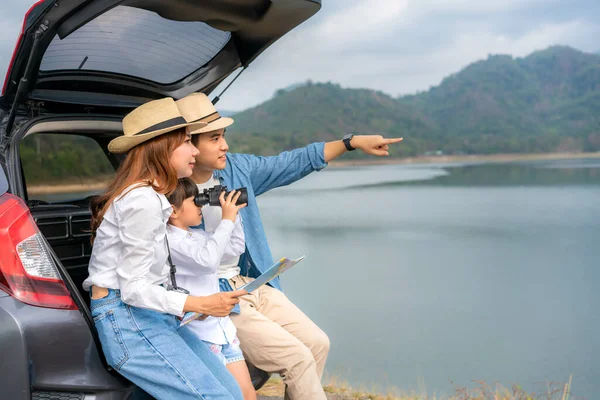 Portrait of Asian family sitting in car with father pointing to view and mother holding maps with daughter looking beautiful landscape through binoculars while vacation together in holiday. Happy family time.