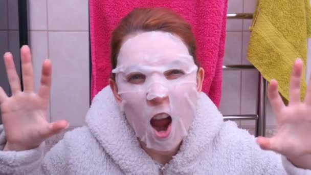 A mulher com uma máscara cosmética no rosto assusta-se. Diversão com uma máscara em seu rosto . — Vídeo de Stock
