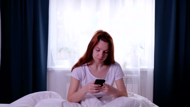 Mujer pelirroja se despierta y se sienta en la cama por la mañana y comienza a usar su teléfono inteligente — Vídeos de Stock