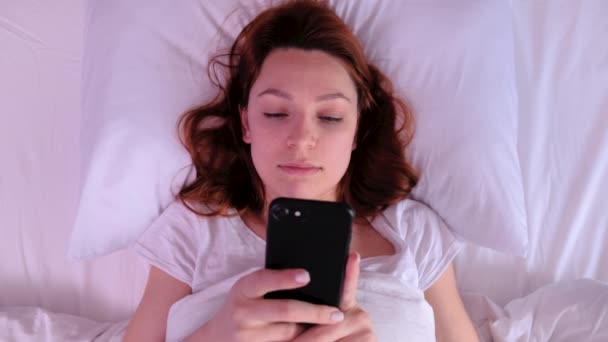 Top view of positive young woman waking up, smiling, stretching in bed and taking her cell phone. Счастливая девушка в белой футболке просыпается утром и начинает пользоваться смартфоном — стоковое видео