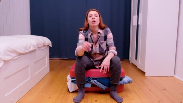 En ung kvinna sitter på en resväska och kan inte stänga den.. — Stockvideo