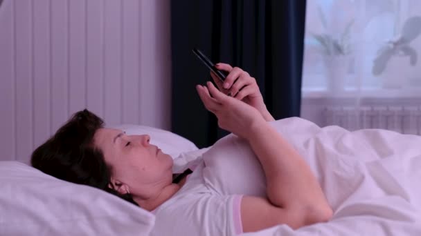 Volwassen vrouw liggend op het bed in de ochtend en met behulp van haar smartphone. — Stockvideo