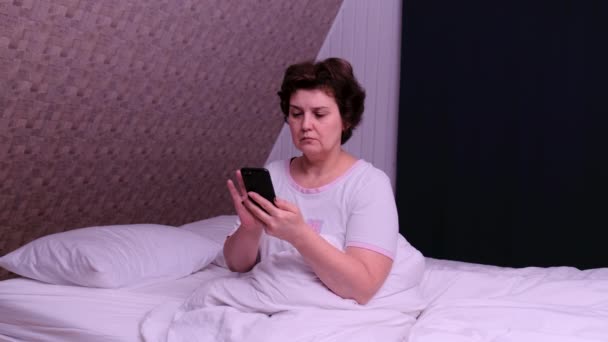 Yetişkin bir kadın yatakta oturur ve uyuduktan sonra telefonu kullanır.. — Stok video