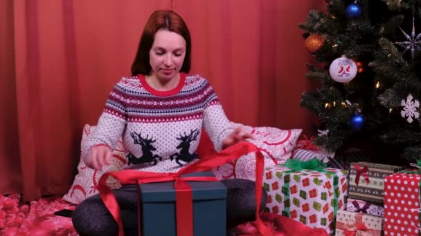 Une femme s'assoit au sapin de Noël et ouvre un cadeau. Elle est très heureuse du cadeau. concept de vacances et nouvelle année . — Video