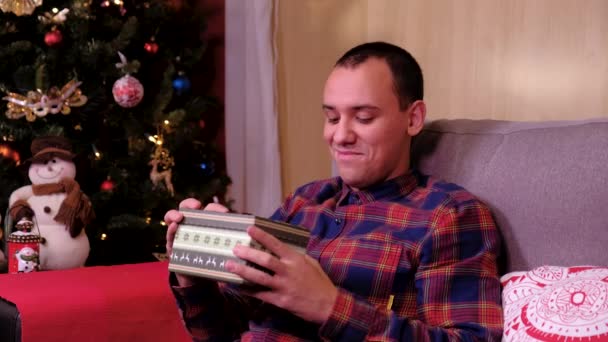 Χριστουγεννιάτικο δώρο - λυπημένος άνθρωπος που βρίσκεται σε έναν καναπέ κοντά στο χριστουγεννιάτικο δέντρο, άνοιγμα δώρο απογοητευμένος και δυστυχισμένος. έννοια των διακοπών και το νέο έτος. — Αρχείο Βίντεο
