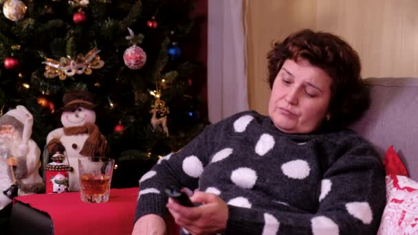 Porträt einer einsamen Frau, die auf dem Sofa neben dem Weihnachtsbaum sitzt und fernsieht. Konzept von Feiertagen und Neujahr. — Stockvideo
