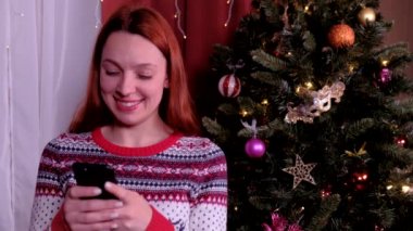 Kadının yanına dekore edilmiş Noel ağacı. Güzel kadın yeni yılda mobil alıcı Tebrikler kullanarak. Arkadaşlarınıza ve ailenize mutlu Noeller dileğiyle