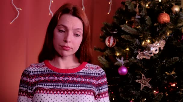 A young woman with red hair looks at the camera and is sad at Christmas. Christmas tree in the background. Concept of holidays, christmas and new year. — Stock Video