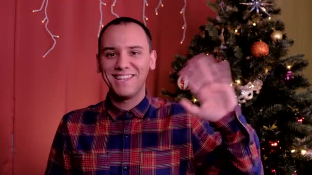 男はカメラを見てクリスマスの挨拶を言う。背景にクリスマスツリー。休日、クリスマス、新年の概念. — ストック動画
