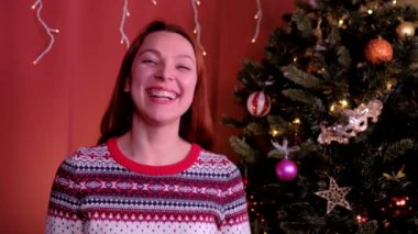 Kızıl saçlı genç bir kadın kameraya bakar ve Noel 'de güler. Arka planda Noel ağacı var. Tatil, Noel ve Yeni Yıl kavramı.