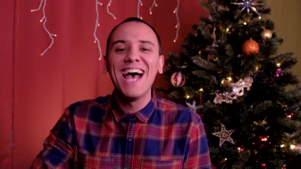 한 남자가 크리스마스에 카메라를 보고 웃는다. 뒤에 크리스마스 트리. 휴일, 크리스마스, 새해의 개념. — 비디오
