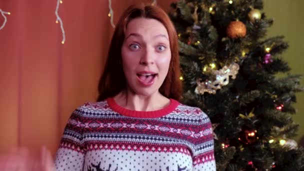 A young woman with red hair looks at the camera and waves a greeting for Christmas. Christmas tree in the background. Concept of holidays, christmas and new year. — Stock Video