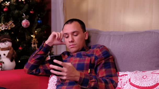 携帯電話でクリスマスツリーの近くに座っている孤独な悲しい男の肖像画。年末年始の概念を — ストック動画