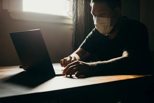 A asian man in medical mask working  on laptop in dark area.
