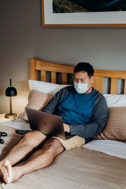 Asyalı adam tıbbi maske takıyordu. Yatak odasında oturup dizüstü bilgisayar kullanmak..
