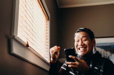 Asyalı bir adam cep telefonu tutuyor ve gülümsüyor. Coronavirus salgınının yol açtığı kısıtlamalar sonucu adam evde kalmaya zorlandı..