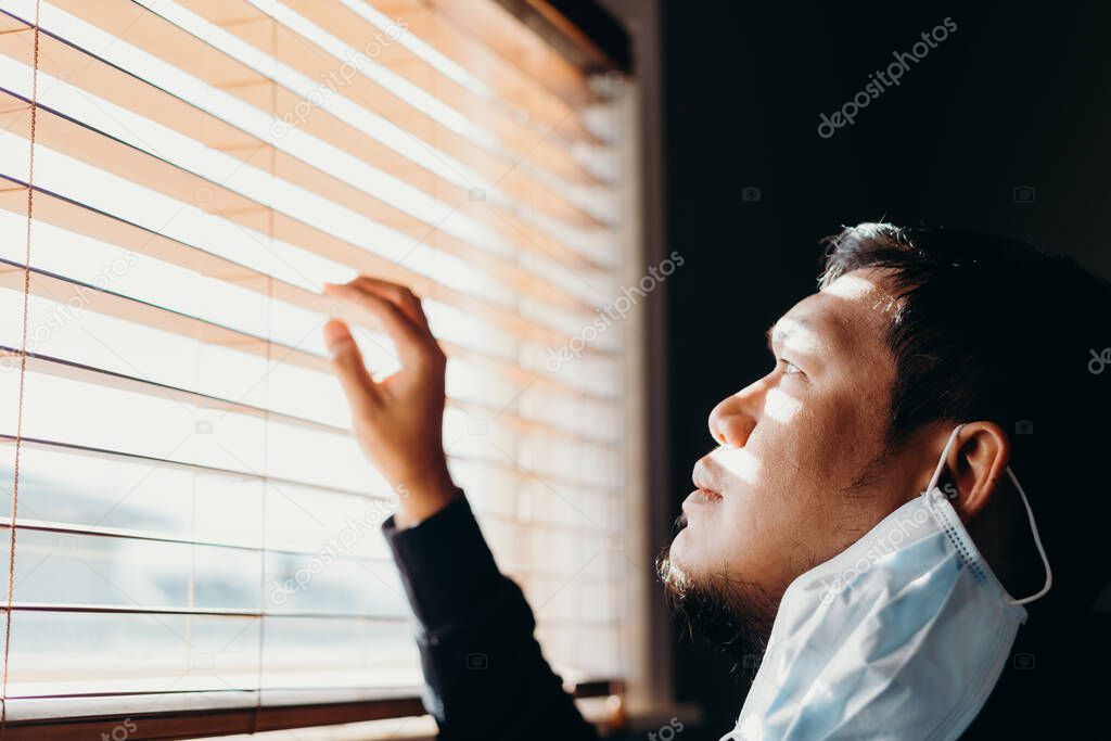 Asian man in isolation looking through window blinds. Man in medical mask forced to stay inside the house as a result of the restrictions caused by the Coronavirus outbreak.